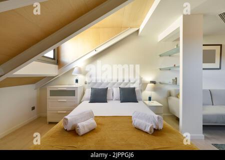 Camera da letto decorata in un appartamento mansardato con soffitti mansardati, testiera rivestita in tessuto bianco capitone, mobili in legno bianco e asciugamani puliti Foto Stock