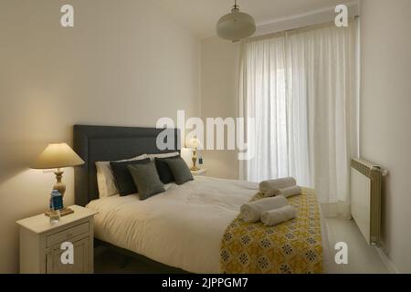 Camera da letto decorata in tonalità tenui, testata imbottita in tessuto blu, cuscini coordinati e lampade doppie, tende bianche e asciugamani puliti arrotolati Foto Stock