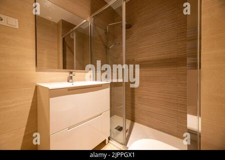 Bagno semplice con cabina doccia, specchio a muro senza cornice, box doccia con porta a vetri scorrevole e armadietto bianco con cassetti Foto Stock