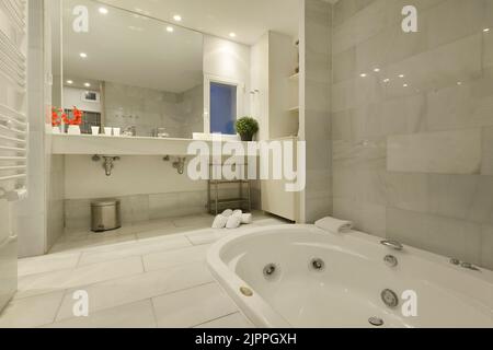 Bagno con specchio in vetro che copre una parete, lungo piano in marmo bianco con due lavandini e vasca idromassaggio sul pavimento Foto Stock