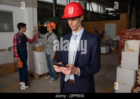 Un ingegnere utilizza un tablet sul cantiere mentre indossa un elmetto e gli operai dietro di lui lavorano Foto Stock
