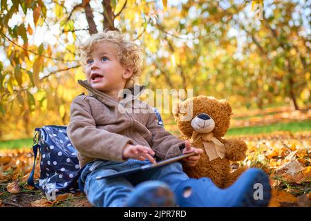 Non lascia mai casa senza il suo orsacchiotto e il suo tablet. Un adorabile ragazzino che usa il tablet mentre si siede all'aperto durante l'autunno. Foto Stock