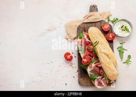 Sandwich baguette con prosciutto, pomodori ciliegini e rucola su un tavolo. Sfondo bianco, vista dall'alto con spazio di copia. Foto Stock