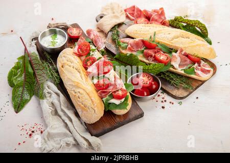 Due panini baguette con salame, mozzarella, lattuga, pomodori e prosciutto su un tagliere. Lunghi sandwich della metropolitana su sfondo bianco. T Foto Stock