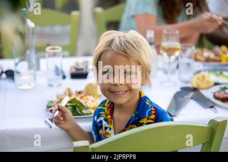 Dolce bambino, seduto nel ristorante estivo all'aperto, mangiare pesce, gamberi, calamari, polpo e patatine fritte Foto Stock