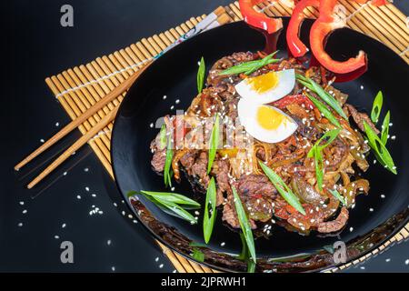 Udon tagliatelle con carne, funghi e verdure. Cosparso di semi di sesamo. Cibo asiatico, arrosto su sfondo nero, spazio copia Foto Stock