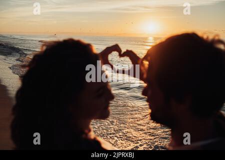 L'uomo e la donna mostrano il cuore con le dita sullo sfondo dell'alba panoramica, giovane coppia che incontra l'alba sulla riva del mare Foto Stock