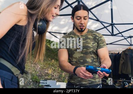 Uomo bianco bearded in camo t-shirt con pistola da mostra a una cliente femminile interessata all'acquisto. Acquistare e vendere armi da fuoco argomento. Ripresa orizzontale per esterni. Foto di alta qualità Foto Stock