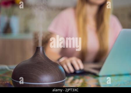 Diffusore di olio essenziale sul tavolo che fuma mentre la donna usa un computer portatile Foto Stock