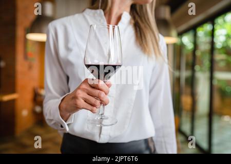 primo piano sulla parte centrale della donna caucasica sconosciuta tenere un bicchiere di vino rosso mentre in piedi indoor indossare bianco camicia copia spazio Foto Stock