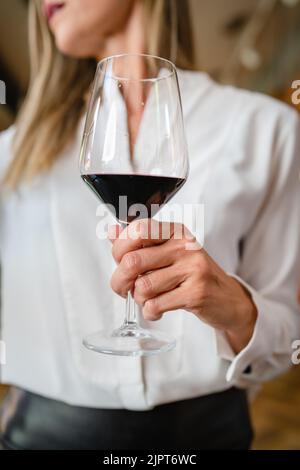 primo piano sulla parte centrale della donna caucasica sconosciuta tenere un bicchiere di vino rosso mentre in piedi indoor indossare bianco camicia copia spazio Foto Stock