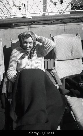 1950s, storica, una donna che indossa un maglione e una sciarpa che si siede all'esterno su un lettino di legno sorridendo per la sua foto. Coprisedolo Woollen coprisedale gambe, Cunard White Star Line, Foto Stock