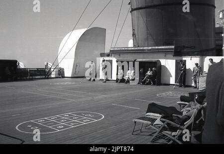 1950s, storico, passeggeri su una nave a vapore, alcuni tenendo pali lunghi, giocare un gioco di ponte, shuffle-board, altri viaggiatori seduti in piccoli rifugi coperti guardare il gioco. Sul lato del ponte, i passeggeri sdraiati su sedie a sdraio in legno con coperte per sedie ricamati cunard. Foto Stock