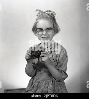 1963, storica, in uno studio fotografico, una giovane ragazza, in posa per la sua foto, negli occhiali e collana delle sue mamme e con un bel fiocco a nastro nei suoi capelli, tenendo una cinepresa dell'epoca, Stockport, Inghilterra, Regno Unito. Foto Stock