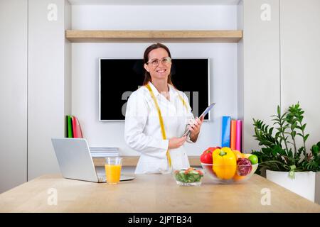 Ritratto di una giovane donna sorridente medico nutrizionista in piedi tenendo bilancia e metro a nastro in sala ufficio moderna e luminosa Foto Stock