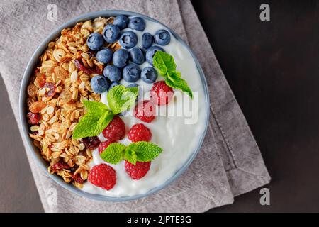 Colazione sana con muesli fatti in casa, yogurt, mirtillo e lampone. Yogurt greco con muesli, frutti di bosco e menta in una ciotola grigia. Vista dall'alto. Copia Foto Stock