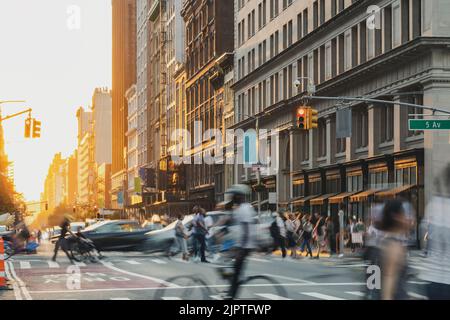 Folle di persone che camminano lungo la strada in un vivace incrocio sulla 5th Avenue a New York City con la luce del tramonto che splende sullo sfondo Foto Stock