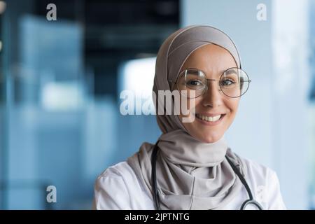Primo piano ritratto fotografico di giovane bella donna musulmana medico, infermiera in occhiali sorridente e guardando la macchina fotografica, donna araba che lavora in clinica moderna ufficio indossare hijab e abito medico Foto Stock