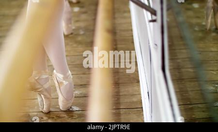 Primo piano, nella sala da ballo, giovani ballerine in leotards viola eseguire pas de bourre suivi in quinta posizione, in piedi sui piedi in scarpe pointe vicino a barre a specchio in classe balletto... Foto di alta qualità Foto Stock