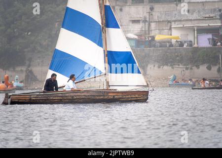 piccola barca a vela colorata con persone sedute su di essa passando barche a file e pedalò in una giornata di nebbia con edifici sullo sfondo sul lago di Foto Stock