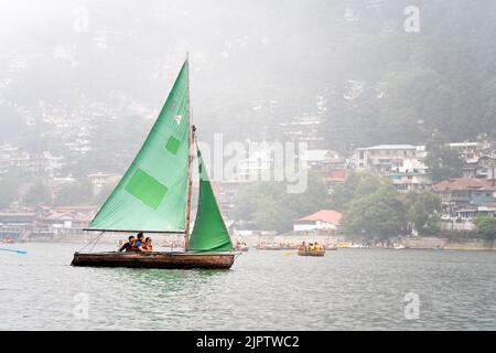 piccola barca a vela colorata con persone sedute su di essa passando barche a file e pedalò in una giornata di nebbia con edifici sullo sfondo sul lago di Foto Stock