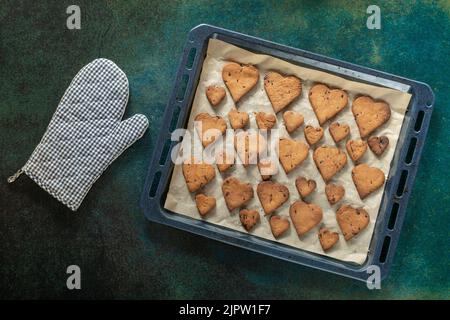 Biscotti a forma di cuore con gocce di cioccolato su una teglia da forno vicino a un guanto da forno. Foto Stock