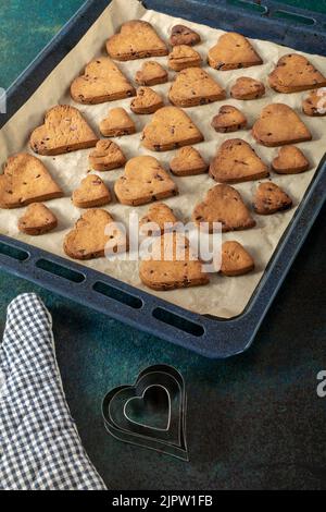 Biscotti a forma di cuore con gocce di cioccolato su una teglia da forno vicino a un guanto da forno e forme di torta. Foto Stock