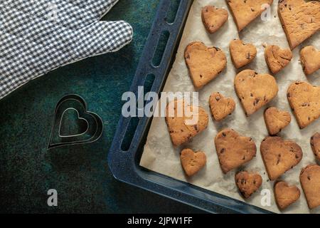 Biscotti a forma di cuore con gocce di cioccolato su una teglia da forno vicino a un guanto da forno e forme di torta. Foto Stock