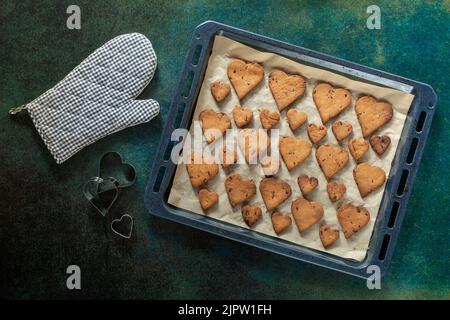 Biscotti a forma di cuore con gocce di cioccolato su una teglia da forno vicino a un guanto da forno e forme di torta. Foto Stock