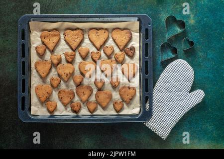 Biscotti a forma di cuore con gocce di cioccolato su una teglia da forno vicino a un guanto da forno e forme di torta. Foto Stock