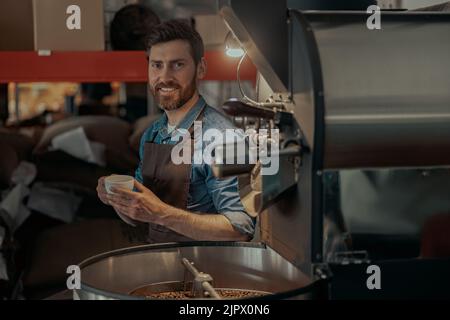 Sorridente imprenditore con una tazza di caffè fresco seduto vicino alla macchina di tostatura in proprio stabilimento Foto Stock