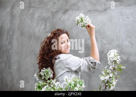 Bella ragazza con lunghi capelli castani ricci in camicia di lino tiene fiorito ramo di albero di mela in mano contro lo sfondo di parete di cemento strutturato. Primavera fioritura tempo. Giornata dell'indipendenza dell'Ucraina Foto Stock