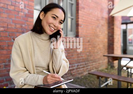 Sorridente giovane imprenditore asiatico parlando sul cellulare seduto all'aperto. Foto Stock