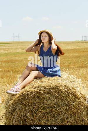 Una bella ragazza in un cappello di paglia siede su un rotolo di fieno e guarda sognatamente il cielo Foto Stock