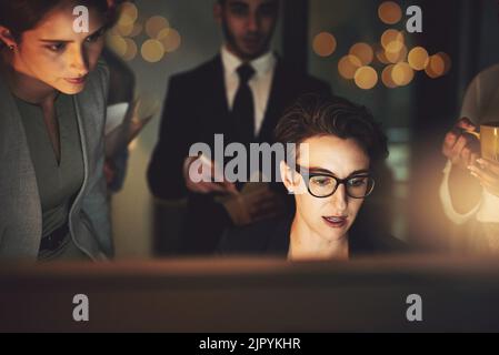 Lavorare sotto pressione è un lavoro che faccio bene: Una giovane donna che lavora tardi con colleghi in piedi in background. Foto Stock