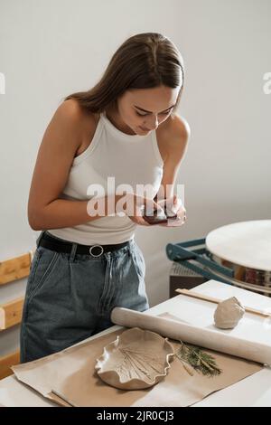 Foto di ceramiche fatte a mano in officina. Ceramica blog proprietario fare foto di lastra di argilla su smartphone lavorando in studio. Donna blogger artista Foto Stock