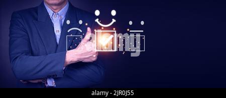 Concetto di valutazione del servizio clienti. Il maschio asiatico sorridente sta usando uno smartphone e sta premendo il sorriso del simbolo del volto in soddisfazione sul touch virtuale sc Foto Stock