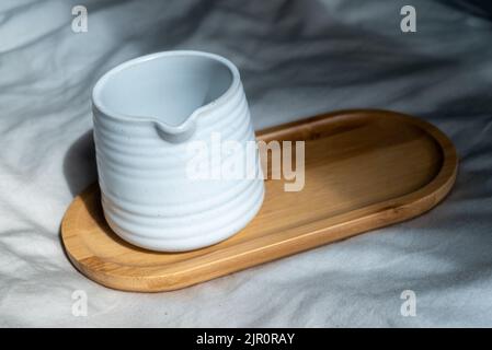 Tazza di argilla bianca o tazza di ceramica sulla piastra di legno. Soggetto su sfondo di stoffa e luce naturale. Foto Stock