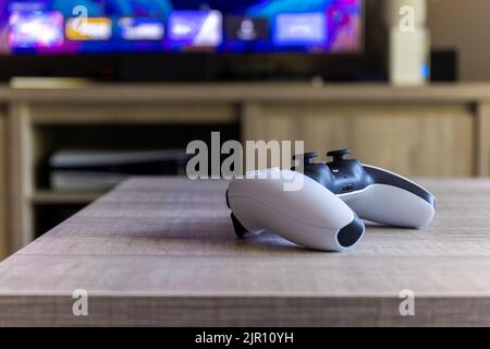 BRECHT, BELGIO – AGOSTO 14 2022: Un ritratto laterale di un controller wireless Sony PlayStation 5 su un tavolo di legno di fronte a un televisore che mostra la P Foto Stock