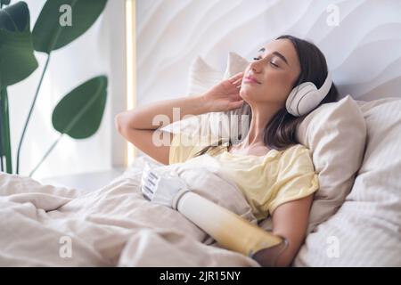 Ragazza felice con braccio bionico rilassante ascoltare musica cuffie a letto a casa. Foto Stock