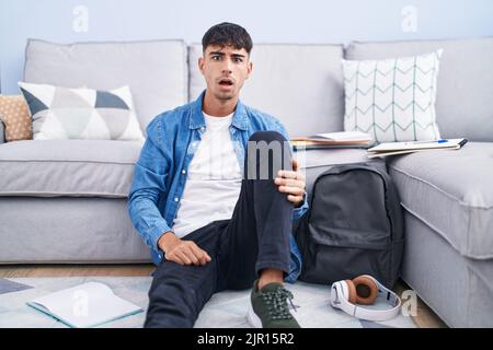 Giovane uomo ispanico seduto sul pavimento che studia per l'università in faccia d'urto, che guarda scettico e sarcastico, sorpreso con la bocca aperta Foto Stock