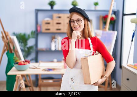 Giovane donna rossa in studio d'arte che tiene custodia d'arte guardando fiducioso la fotocamera sorridendo con braccia incrociate e mano sollevata sul mento. Pensiero positiv Foto Stock