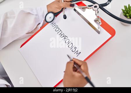 Donna afroamericana che indossa medico uniforme scrittura ipotiroidismo parola sulla clipboard in clinica Foto Stock
