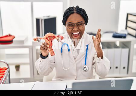 Donna afroamericana medico che tiene modello anatomico di organo genitale femminile celebrando la vittoria con sorriso felice e vincitrice espressione con sollevato Foto Stock
