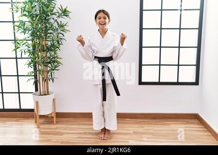 Giovane ragazza ispanica che indossa kimono karate e cintura nera festeggia sorpreso e stupito per il successo con le braccia sollevate e gli occhi aperti. Concetto vincitore. Foto Stock