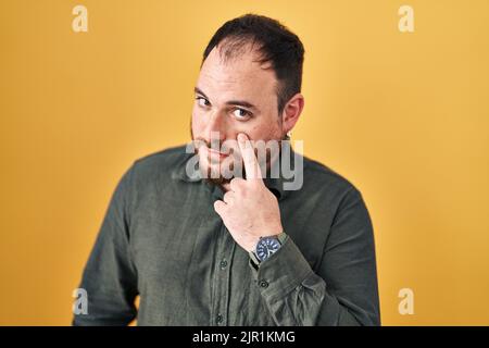 Più taglia uomo ispanico con barba in piedi su sfondo giallo puntando all'occhio che ti guarda gesto, espressione sospetta Foto Stock