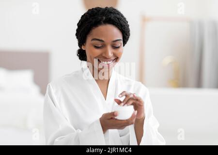 Cosmetici per la cura della pelle. Donna nera sorridente cercando nuova crema idratante a casa Foto Stock