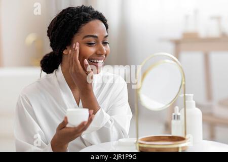 Anti-invecchiamento Skincare. Ritratto di sorridente attraente nero femmina applicazione idratante viso Crema Foto Stock