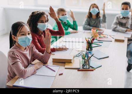 Bambini della scuola multiculturale che indossano maschere facciali che alzano le braccia in aula Foto Stock