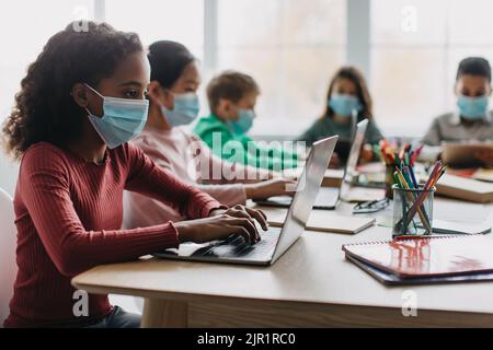 Diversi ragazzi scolastici che utilizzano computer che indossano maschere facciali in aula Foto Stock